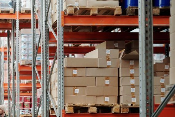 gallery-02 Two workers handle a package in a spacious warehouse surrounded by shelves stocked with boxes and products.