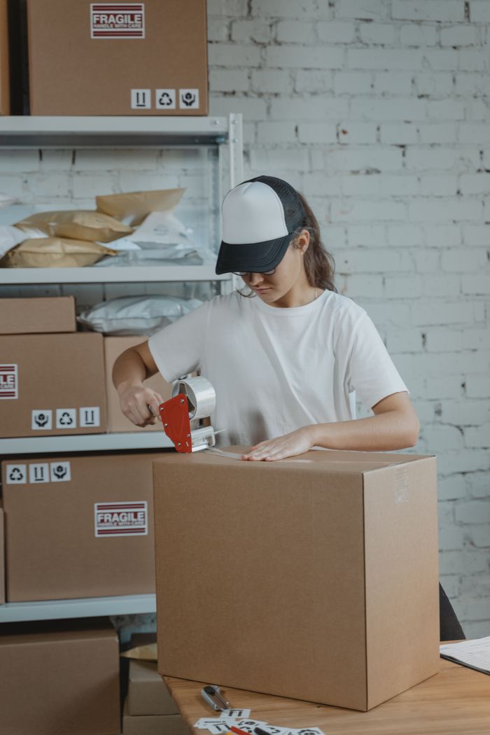 cta-02 Young female worker sealing cardboard boxes in an indoor warehouse setting.