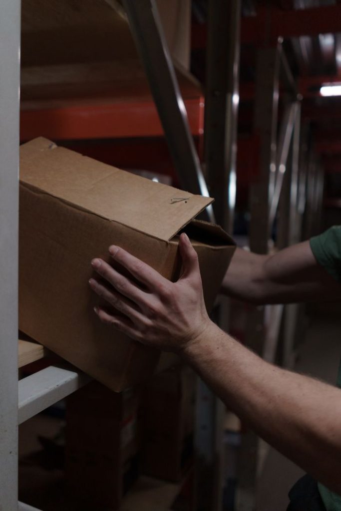 pexels-photo-7019212-7019212 Close-up of a worker placing a cardboard box on a shelf in a warehouse setting.